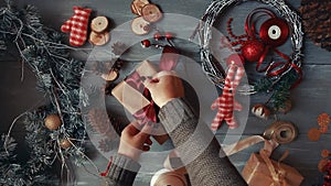 A top-down plan. Fully visible the table with the decorations. Female hands put and finalize Christmas gift wrapped in