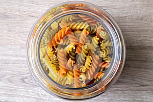 A top down portrait of a glass pot, jar or bowl full of spirelli in three different colors. The uncooked tricolore italian pasta
