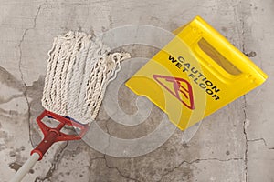 Top down shot of a yellow caution wet floor sign beside a mop on a freshly cleaned concrete surface, signaling safety