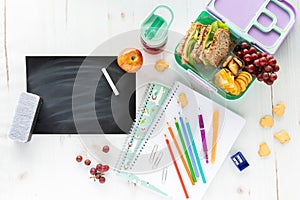 Top down view of an assortment of school supplies and lunch along with a blank chalkboard, chalk and eraser.