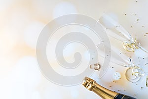 A top down view of champagne flutes and bottle as well as wedding rings, against a bokeh background and glitter.
