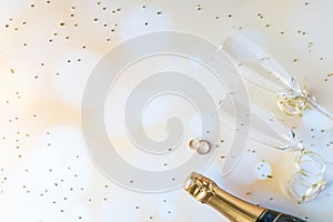 A top down view of champagne flutes, bottle and wedding bands against a bokeh background and glitter all around.