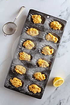 Top down view of raw madeleine cake dough in a baking tin, diagonal, with sieve of flour and lemon next to it