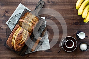 A top down view of a sliced banana bread on a wooden board with a cup of coffee and cream and sugar.