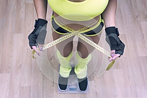 Top-down view to fitness woman standing on scale and taking waist measurements with metric tape. Concept of losing