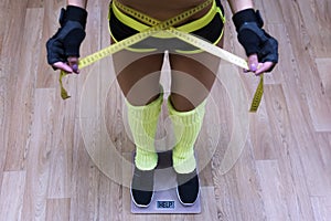 Top-down view to fitness woman standing on scale and taking waist measurements with metric tape. Concept of losing weight in gym.