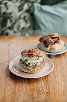 Top-down view of two plates of burger bagel breakfast sandwiches, with eggs, chicken, sauce, and vegan on wooden table