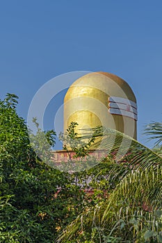 Top of a golden color Lord Shiva Lingam temple in Puttaparthi with green trees