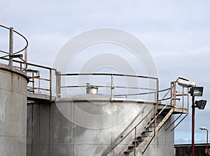 Top of large old silver steel industrial storage tanks with control valves rusting stairs and walkway against a blue sky