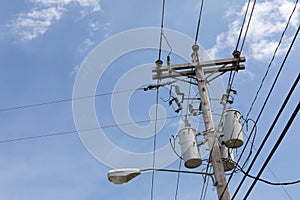 Top of a power pole with multiple transformers, insulators, wires, and a street light against a blue sky with clouds, creative cop