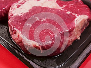 Top quality rib eye steak on a black plastic tray and red color surface background. Premium meat cut product. Butcher craft. Food