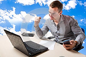 On top of success concept. Blue sky on background. Young businessman sitting at his desk, celebrating success with arms raised