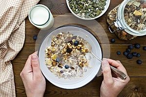Top vie of hand holding spoon of oats for breakfast, granola with dried fruit and blueberry, milk and honey. Wooden background.