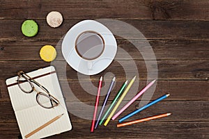 Top viev on a coffee mug, macaroons, colored pencils and a notebook. Wooden background