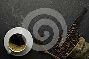 Top view above of Black hot coffee with smoke and milk foam in white ceramic cup with coffee beans roasted in sack bag on dark sto