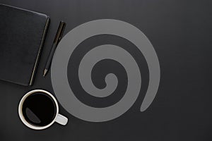 Top view above of dark black office desk table with notebook and coffee cup with a pen.