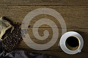 Top view above of Hot fresh black coffee with milk foam in white ceramic cup with coffee beans roasted in sack bag on wood table b