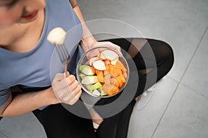 Top view of active woman sitting and eating cut red and green apple mixed with carrot in glass bowl picking with a fork.