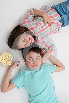 Top view adorable kids, mischievous sister and brother eating pop corn looking at camera, lying over white background.