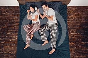 Top view. Adult handsome man plans to wake up with alarm clock sleeping young woman lying on bed.