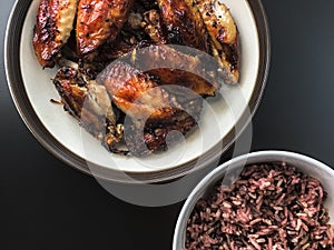 Top view of air fried chicken wings on ceramic dish serve with small bowl of cooked Rice berry. All on black background.