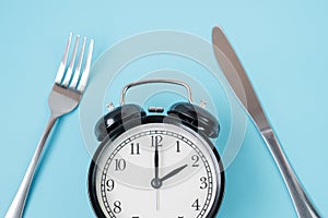 Top view alarm clock with knife and fork on blue background. Intermittent fasting, Ketogenic dieting, weight loss, meal plan and