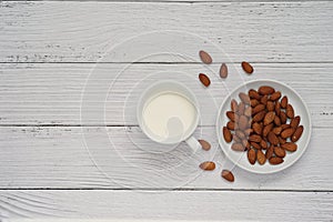 Top view of almonds milk in a white ceramic cup and roasted almonds on white ceramic plate placed on white wooden background.