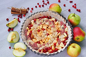 Top view on the apple pie in the baking dish decorated with fresh apples, cranberry, spices, cinnamon sticks on the gray table