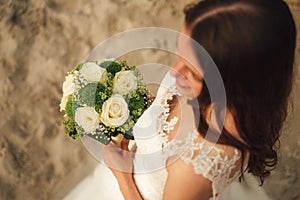 Top view of attractive brunette bride holding wedding bouquet. White roses flowers. Evening light on sandy beach