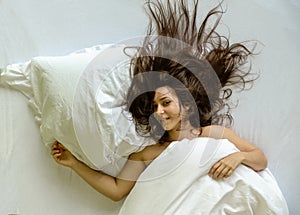 Top view of attractive, young, naked, dark-haired woman, mouth open, laughing, hair wild on the sheets, relaxing in bed, sleeps,