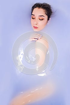 Top view of attractive young woman keeping eyes closed while enjoying luxurious bath. Colored purple water,