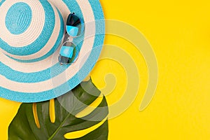 Top view on beach accessories on yellow background - sunglasses, striped blue hat and monstera leaf. Concept of the long