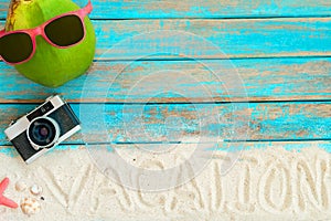 top view of beach sand with coconut, sunglasses, retro camera, starfish and shells on blue wooden background.