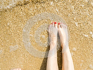 Top view of beautiful female feet with bright red pedicure on the sand of the beach. The sea wave washes women`s feet