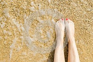 Top view of beautiful female feet with bright red pedicure on the sand of the beach. The sea wave washes women`s feet