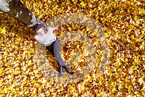 Top view. Beautiful happy young woman sitting under tree and dreaming on the autumn maple leaves in the park. Aerial, drone view.