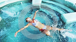 Top of view of beautiful tan young woman in bikini and sunglasses swims in swimming pool in crystal clear water jet pool