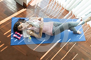 Top view of beautiful woman wearing sportswear looking camera and lying on yoga mat on wooden floor near dumbbells and