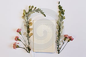 Top view of a bible with white leather cover and flowers on a white surface