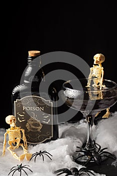 Top view of black Halloween cocktail glass with skeletons and bottle of poison on spider web, black background,
