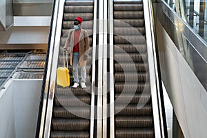 Top view of Black man with yellow luggage standing on escalator at airport terminal wearing face protective mask during virus