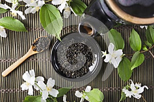 Top view of black teapot and saucer full of dry green tea, wooden spoon and aromatic fresh jasmine flowers on the green wicker mat