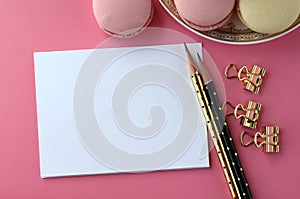 Top view of blank note card, laptop, cell phone and French macarons on pink background. Feminine workspace desktop with copy space