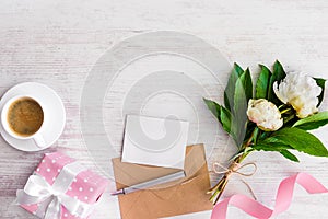 Top view of blank note, kraft envelope, coffee cup and peony flowers over white wood rustic background.Copy space.