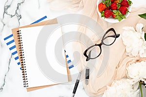 Top view blank paper notepad, pastel beige scarf, bowl of strawberry, ink pen and white peony flowers on marble background.