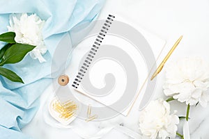 Top view blank paper notepad, white peony flowers, blue scarf, golden clips on marble background. Minimal flat lay style