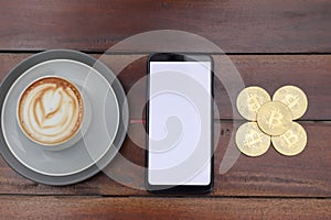 Top view of blank screen mobile phone with a pile of bitcoin and cup of coffee on wooden table. Cryptocurrency trading concept