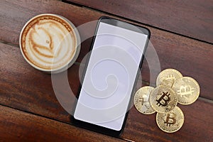 Top view of blank screen mobile phone with a pile of bitcoin and cup of coffee on wooden table. Cryptocurrency trading concept