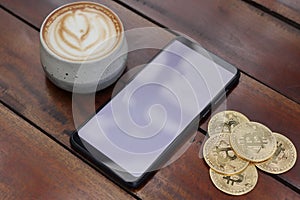 Top view of blank screen mobile phone with a pile of bitcoin and cup of coffee on wooden table. Cryptocurrency trading concept