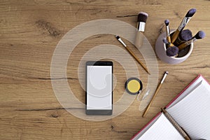 Top view of blank screen of smartphone,makeup tools,brushes, opened notebook on the wooden table.Small business influencer concept